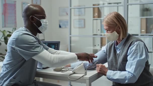 若いアフリカ系アメリカ人男性医師は 病院の職場に座り 診察中に成熟した女性患者の血圧を測定しながら メートル法を使用して均一で保護マスクで — ストック動画