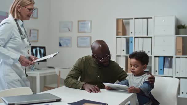有经验的临床医生带来了医疗文件 并将其交给年轻人签字 同时走向他和他可爱的小儿子一边看着平板电脑屏幕 一边看在线视频 — 图库视频影像