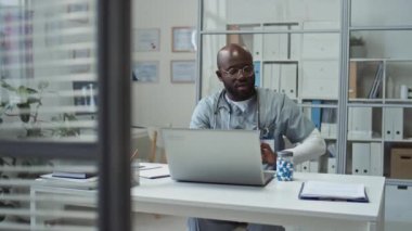 Genç kendinden emin Afrikalı Amerikalı üniformalı doktor kliniklerde dizüstü bilgisayarın önünde oturuyor, hastalara tıbbi tavsiyelerde bulunuyor ve etkili yeni ilaçlar öneriyor.