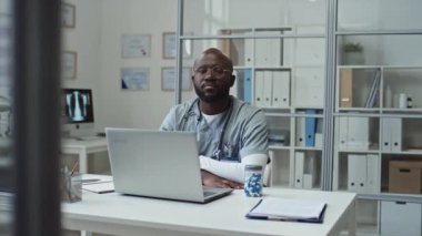 Genç özgüvenli Afrikalı Amerikalı üniformalı doktor bilgisayarının önünde otururken kameraya bakıyor. Masasında tıbbi dokümanlar ve bilgisayar monitörü olan raflara karşı.