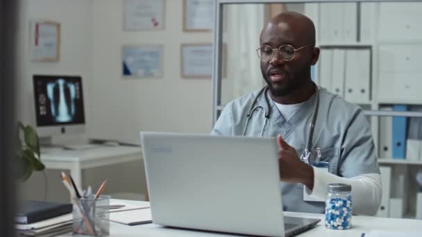 オンライン相談中にノートパソコンの前で職場に座って患者に薬を推薦する制服や眼鏡の若いアフリカ系アメリカ人の一般開業医 — ストック動画