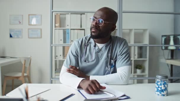 若い深刻なアフリカ系アメリカ人男性の臨床家眼鏡と青い制服彼の職場で医療室に座って カメラを見て — ストック動画