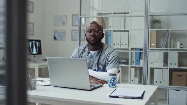 若い自信に満ちたアフリカ系アメリカ人のオンライン医師は 医療文書や机の上のコンピュータモニタで棚に対してラップトップの前に彼の職場で座っている間 カメラを見て均一です — ストック動画