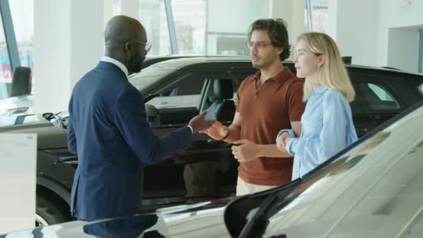 幸せな若いカップルに新しい車のキーを渡すディーラーセンターで働くアフリカ系アメリカ人営業担当者 — ストック動画