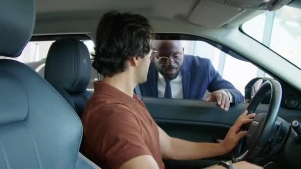 中东年轻人坐在现代汽车的前排 在车行的陈列室里探索汽车的内部 并与非洲裔美国人推销员进行对话 — 图库视频影像