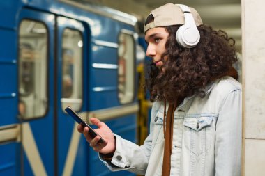 Akıllı telefonlu şık zoomer metroda durup mavi trene bakarken kulaklıkla müzik dinliyor.