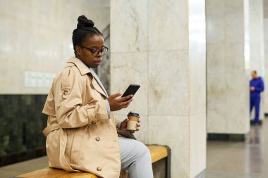 İstasyonda bankta oturan, akıllı telefondan mesaj atan ve metro trenini bekleyen Afro-Amerikalı genç bir kadının yan görüntüsü.