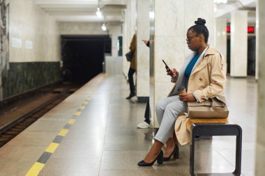 Metro istasyonunda oturup internetten video izlerken ya da mesajlaşırken cep telefonlu şık siyahi bir kadın metro trenini bekliyor.