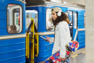 Mavi metro treninin önünde duran ve metroyla seyahat ederken cep telefonu kullanan modern gençlerin yan görüntüsü.