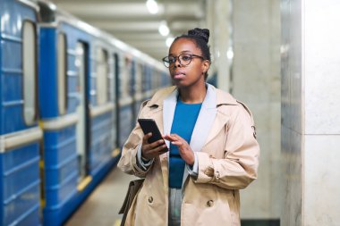 Cep telefonlu genç Afrikalı iş kadını metro istasyonunda mavi metro trenine karşı duruyor ve ileriye bakıyor.