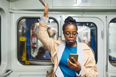 Metroya binerken ve akıllı telefondan video izlerken tırabzanla tutulan gözlüklü, ciddi bir Afrikalı Amerikalı kadın.