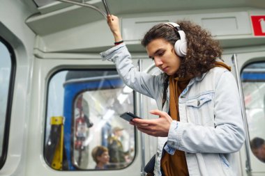 Kulaklık takmış, trende tırabzanla durup internetten film ya da video izlerken akıllı telefon ekranına bakan ciddi bir genç adam.