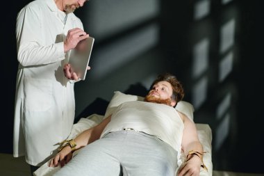 Laboratuvar önlüğü giymiş olgun bir doktor klinik tedavi sırasında yatağa bağlı genç bir erkek hastaya tablet ekranda bir şey gösteriyor.
