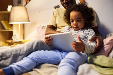 Küçük çocuk yatak odasında babasıyla yatakta otururken dijital tablette çizgi film izliyor.