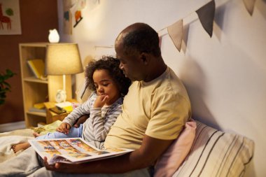 Afrikalı Amerikalı baba çocuğuna masal okuyor. Yatakta oturup kitaptaki resimleri izliyorlar.
