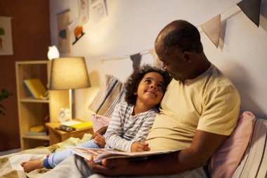 Afrikalı Amerikalı baba, onu yatakta kucaklarken uyumadan önce çocuğa kitap okuyor.