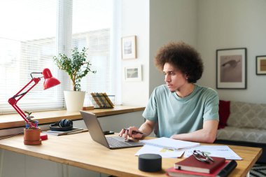 Genç adam masada belgelerle ve diğer aletlerle otururken dizüstü bilgisayar kullanıyor.