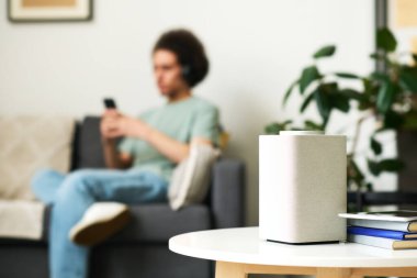 Arka planda akıllı telefon kullanan bir adamla odadaki sehpanın üzerinde duran akıllı hoparlörün yakın plan görüntüsü.