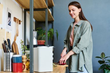 Gülümseyen genç kadın mutfakta yemek hazırlıyor ve akıllı hoparlörden müzik dinliyor.