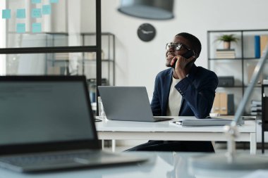 Açık Uzay Ofisi 'nde bilgisayarın önünde çalışırken akıllı telefondan müşteriyle konuşan mutlu genç Afrikalı Amerikalı simsar.