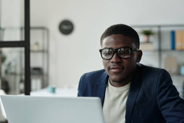 Genç, beyaz yakalı, gözlüklü, dizüstü bilgisayarın önünde oturmuş görüntülü sohbet sırasında ekrana bakıyor.