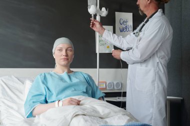 Onkolojik hastalığı olan genç bir kadın hasta yatağında otururken laboratuvar önlüklü erkek onkolog yanında duruyor ve damlatıcıyı hazırlıyor.