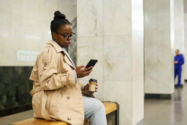侧面看年轻的非洲裔美国女人 一杯咖啡坐在车站的长椅上 用智能手机发短信 等待地铁列车 — 图库照片