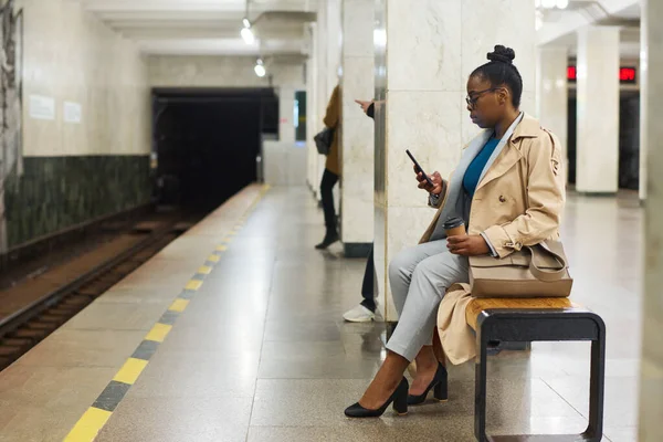 年轻时尚的黑人女性 坐在地铁站边等地铁列车 一边发短信或观看网上视频 — 图库照片