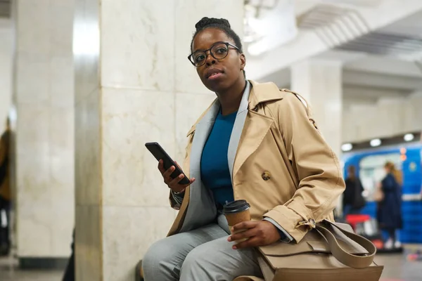 身穿时髦米黄色风衣的年轻非洲裔美国女人坐在地铁站等火车 一边用手机发短信 — 图库照片
