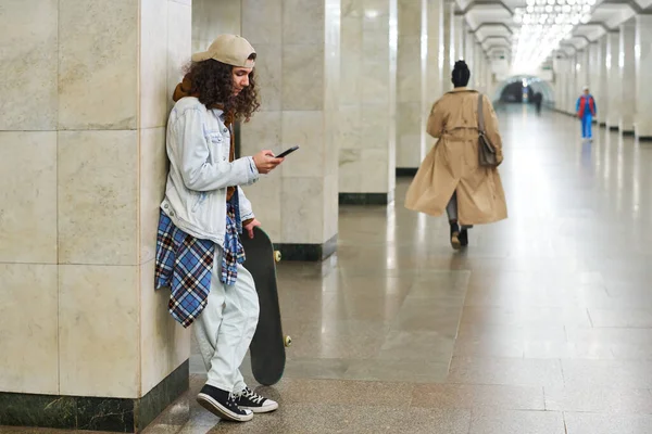 スタイリッシュなティーンスケートボーダーは バックグラウンドで女性と地下鉄駅で大理石の列に対して立っている間 携帯電話でテキストメッセージ — ストック写真