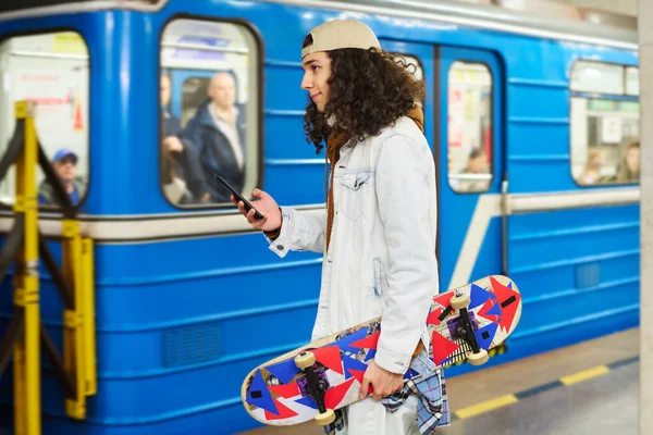 青い地下鉄の列車の前に立っているスケートボードとスマートフォンを持つ10代の男は プラットフォーム上で停止する前に駅に沿って移動 — ストック写真