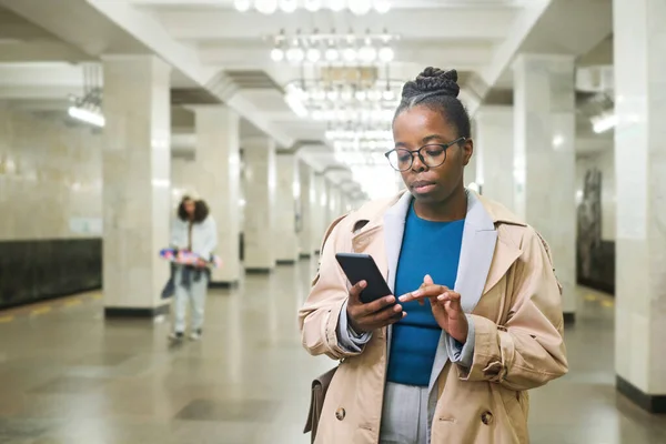 身穿风衣的年轻而严肃的黑人妇女站在地铁站旁边 靠着有柱子的隧道 一边用智能手机通过位置图 — 图库照片