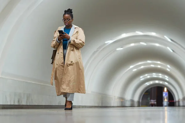 身穿米黄色风衣的年轻非洲裔美国女人一边用智能手机发短信 一边沿着宽阔而长长的地铁隧道走到站台上 — 图库照片