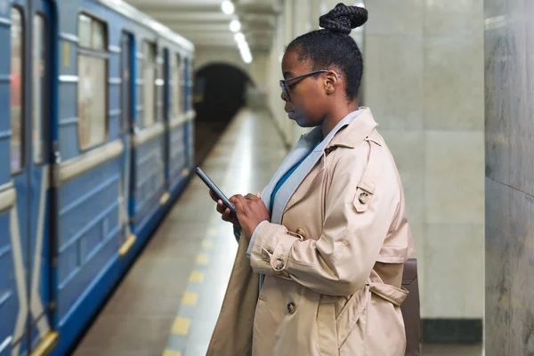 stock image Side view of young black woman scrolling in smartphone or watching video while standing in front of moving blue train at subway station