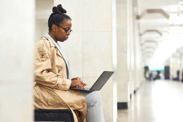 坐在地铁站的长椅上等火车时穿着便服在笔记本电脑键盘上打字的年轻而严肃的女人的侧影 — 图库照片