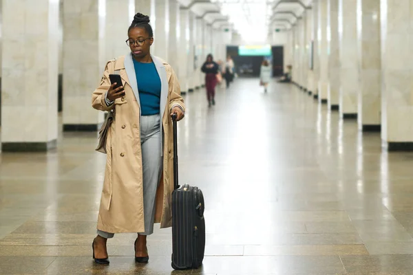 年轻的女旅行者拿着手提箱用智能手机发短信 站在长长的大厅里的摄像机前 靠着有柱子的地铁隧道 — 图库照片