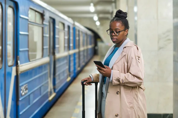 年轻而严肃的女商务旅行者 背着手提箱用智能手机发短信 站在镜头前 背对着移动的火车 — 图库照片
