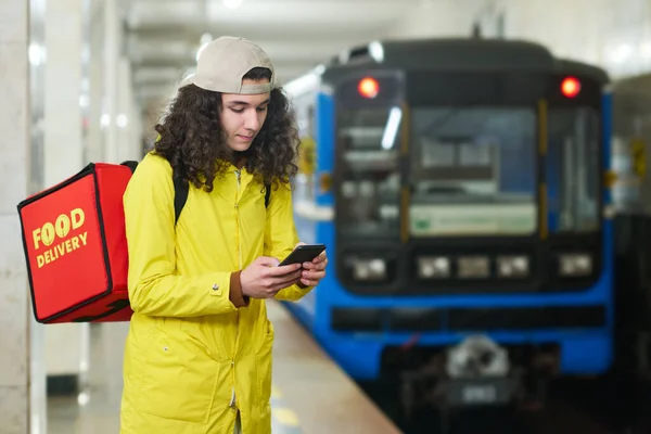 穿着黄色制服背着红色大背包的家伙用智能手机站在地铁站的摄像机前与火车抗衡 — 图库照片