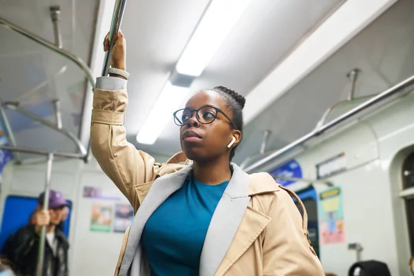 年轻的黑衣女子穿着便服 戴着眼镜 一边用耳机听着音乐 一边手牵着扶手在乘地铁上班的路上 — 图库照片