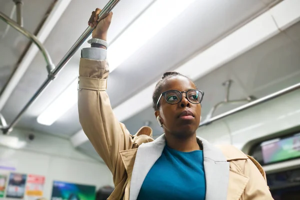 年轻而严肃的非洲裔美国女人 戴着耳机 手握扶手 坐地铁时透过眼镜向前看 — 图库照片