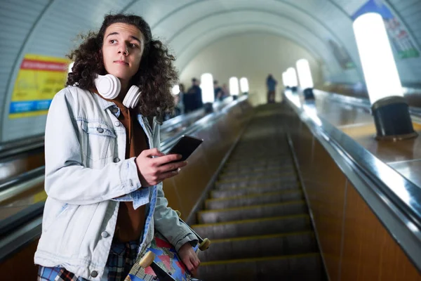 Подростковый Скейтбордист Смартфоном Стоящим Движущемся Эскалаторе Смотрящим Сторону Длинного Туннеля — стоковое фото