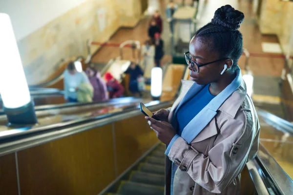 乘坐眼镜的年轻黑人妇女在地铁的自动扶梯上观看智能手机在线视频的侧视图 — 图库照片