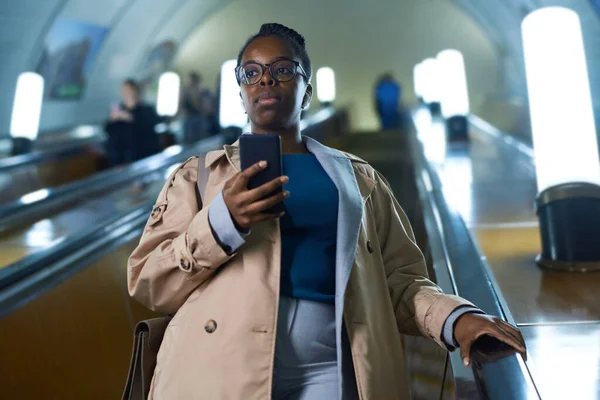 年轻的女通勤者戴着眼镜 穿着便服 在自动扶梯上向前望去 同时靠着地铁内部 并使用智能手机 — 图库照片