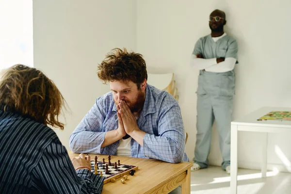 年轻而紧张的男人 一边与其他男性精神病患者玩耍 一边用手捂住脸 看着棋盘 — 图库照片