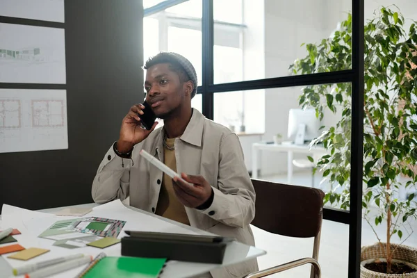 年轻自信的黑人男子穿着便服 坐在写字台旁与同事通电话 桌上有草图 目录和办公用品 — 图库照片