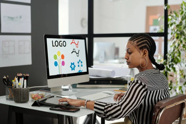 年轻而严肃的非洲裔美国女性网页设计师坐在电脑显示器前使用平板电脑的侧视图 — 图库照片