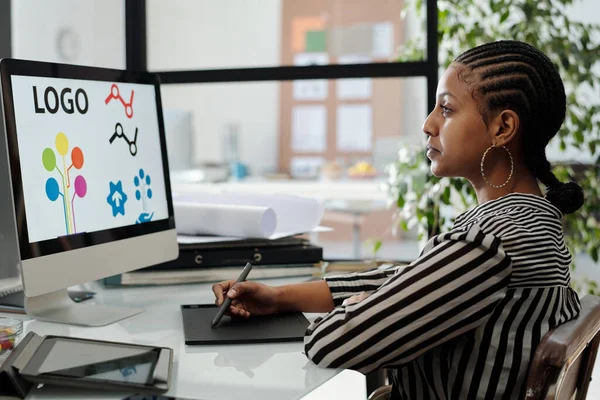 严肃的非洲裔美国女性网络设计师坐在电脑屏幕上印有标识的显示器前 正在研究新项目 — 图库照片