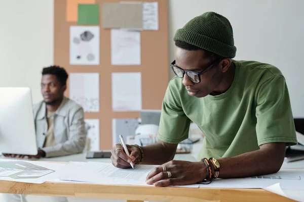 穿着便服和眼镜的年轻的非洲裔美国设计师或建筑师 一边在办公室里画新建筑的草图 一边弯腰在纸上画 — 图库照片
