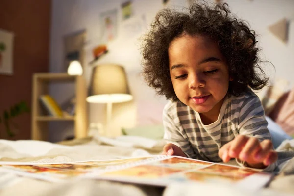 アフリカ系アメリカ人の男の子見る写真で本で寝る前にベッドルームに横たわっている — ストック写真