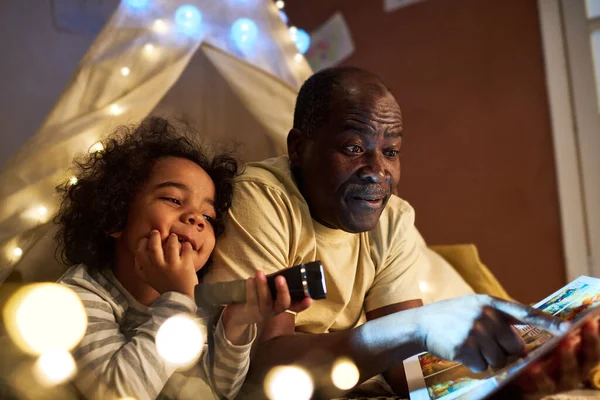 African American dad reading fairytale book to his child while they sitting in tent and using flashlight